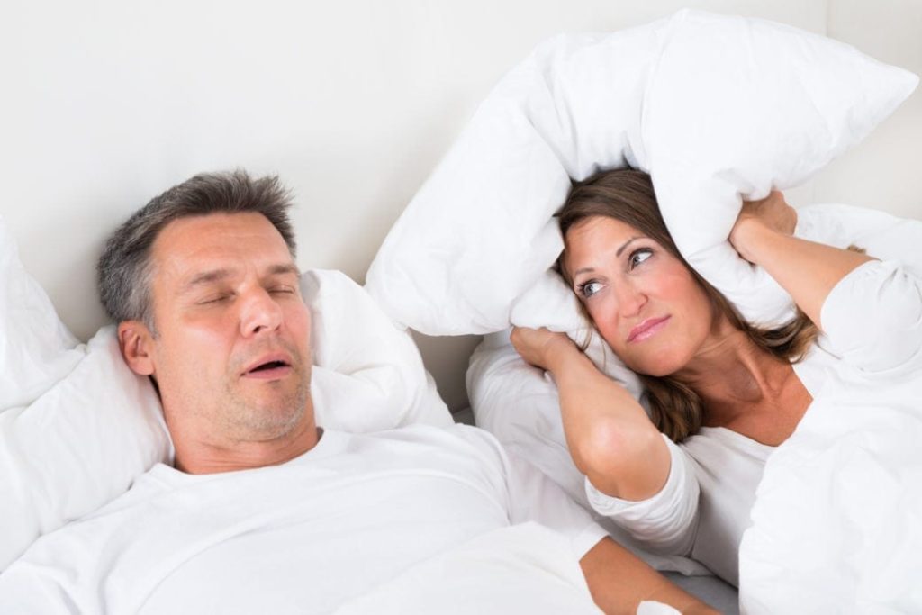 Woman Covering Her Ears With Pillow While Man Snoring suffering from sleep apnea. 